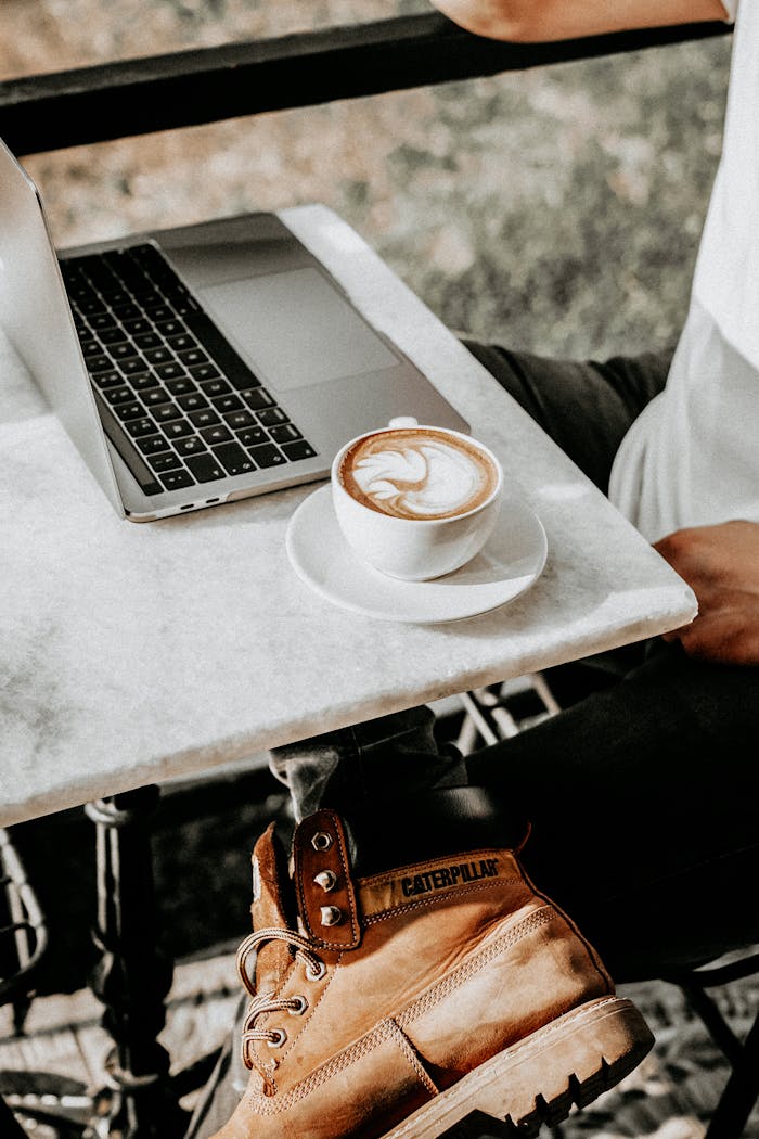 Casual outdoor setting with a man enjoying coffee, working on a laptop with stylish boots.