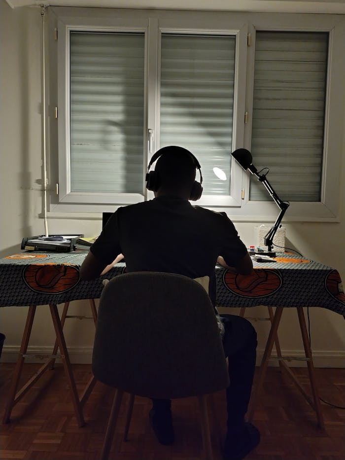 Silhouette of a person working at a desk with headphones, lit by a desk lamp.