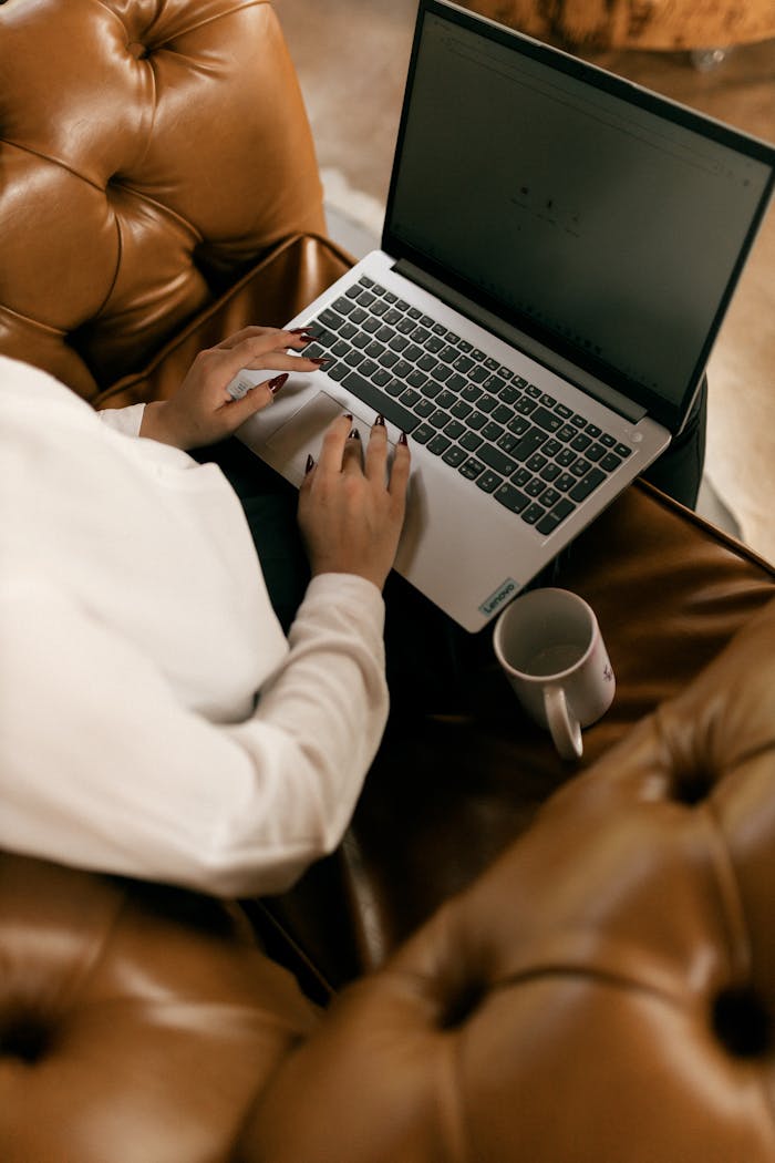 A woman typing on a laptop while sitting on a brown leather sofa with a mug nearby.