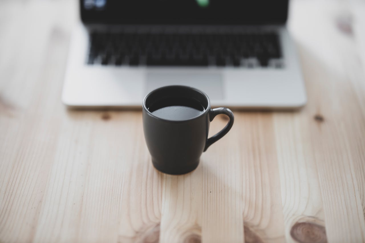 Black coffee mug on wooden table with blurred laptop background. Perfect for work-from-home themes.